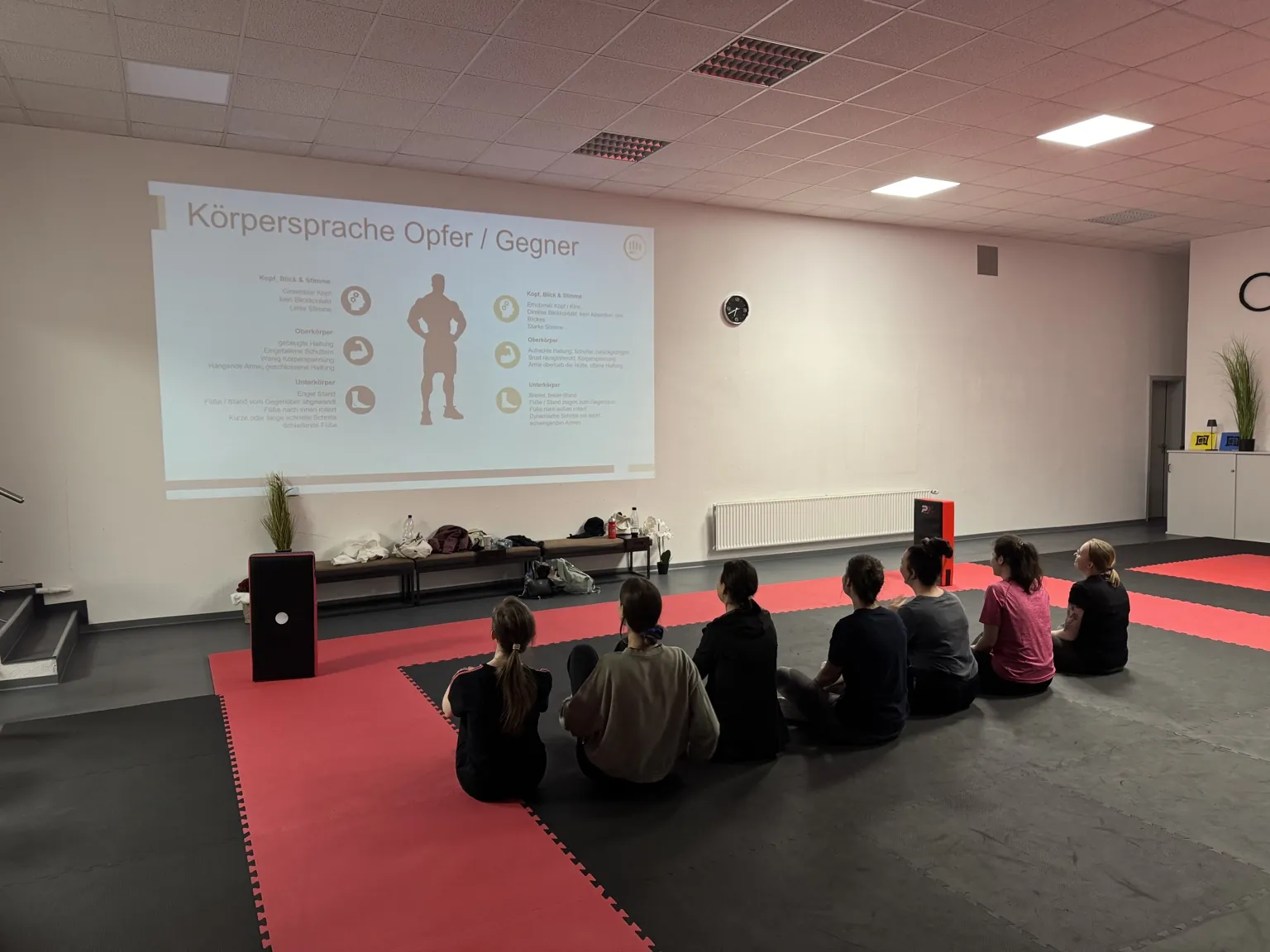 Teilnehmende eines Selbstverteidigungsworkshops sitzen im CONFIDENSE-Studio vor einer projizierten Präsentation zur Körpersprache.