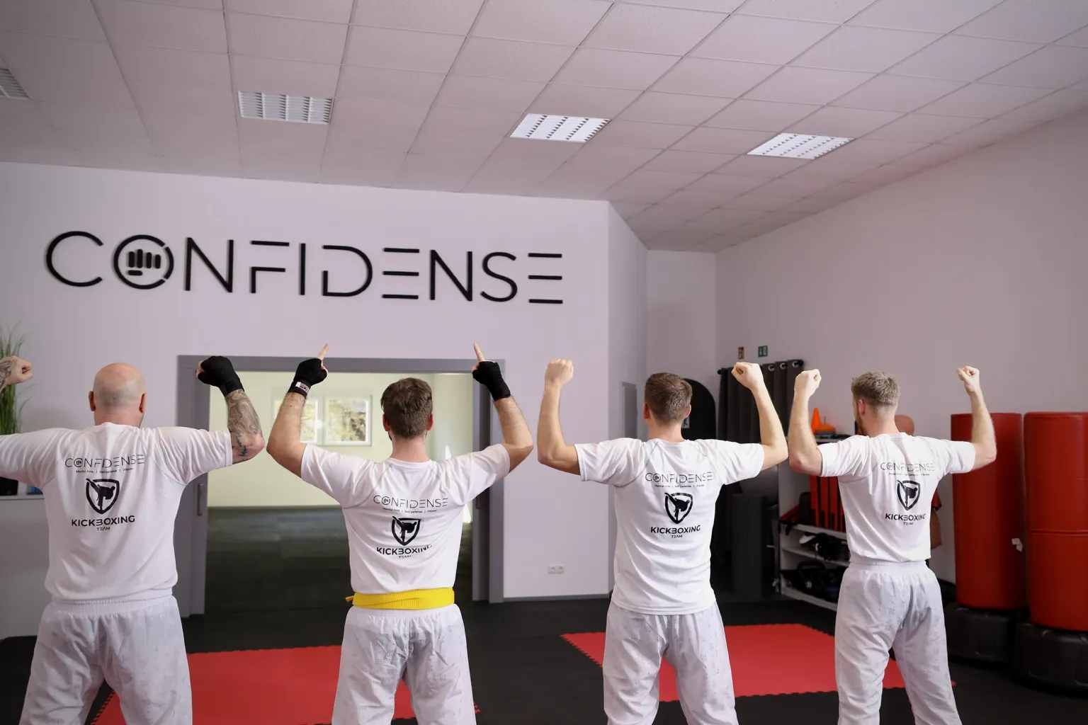 Vier Kickboxer stehen mit dem Rücken zur Kamera im Studio und heben die Arme unter dem großen CONFIDENSE-Schriftzug.