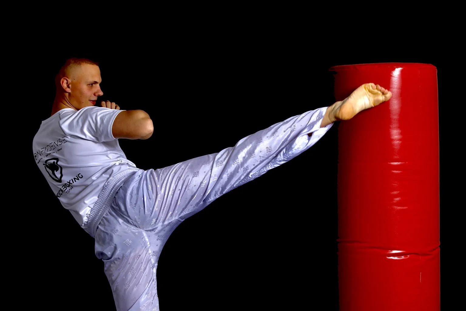Kickboxer in weißer Trainingskleidung führt einen hohen Tritt gegen ein rotes Schlagpolster aus.
