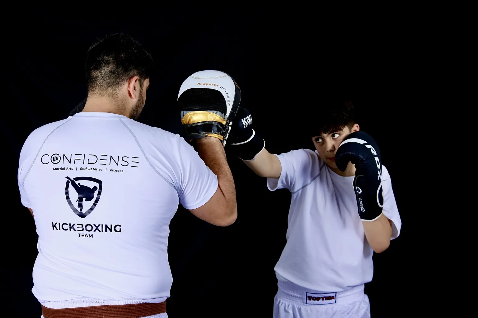 Junger Kickboxer trainiert Schlagtechniken an Pratzen vor schwarzem Hintergrund.