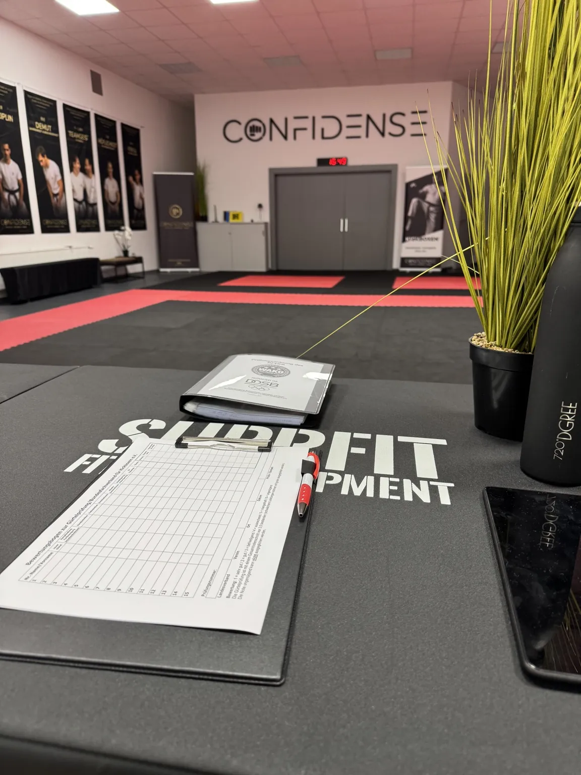 Blick vom Empfang über die Trainingsfläche des CONFIDENSE-Studios mit schwarzer und roter Mattenfläche und großem Wandschriftzug.