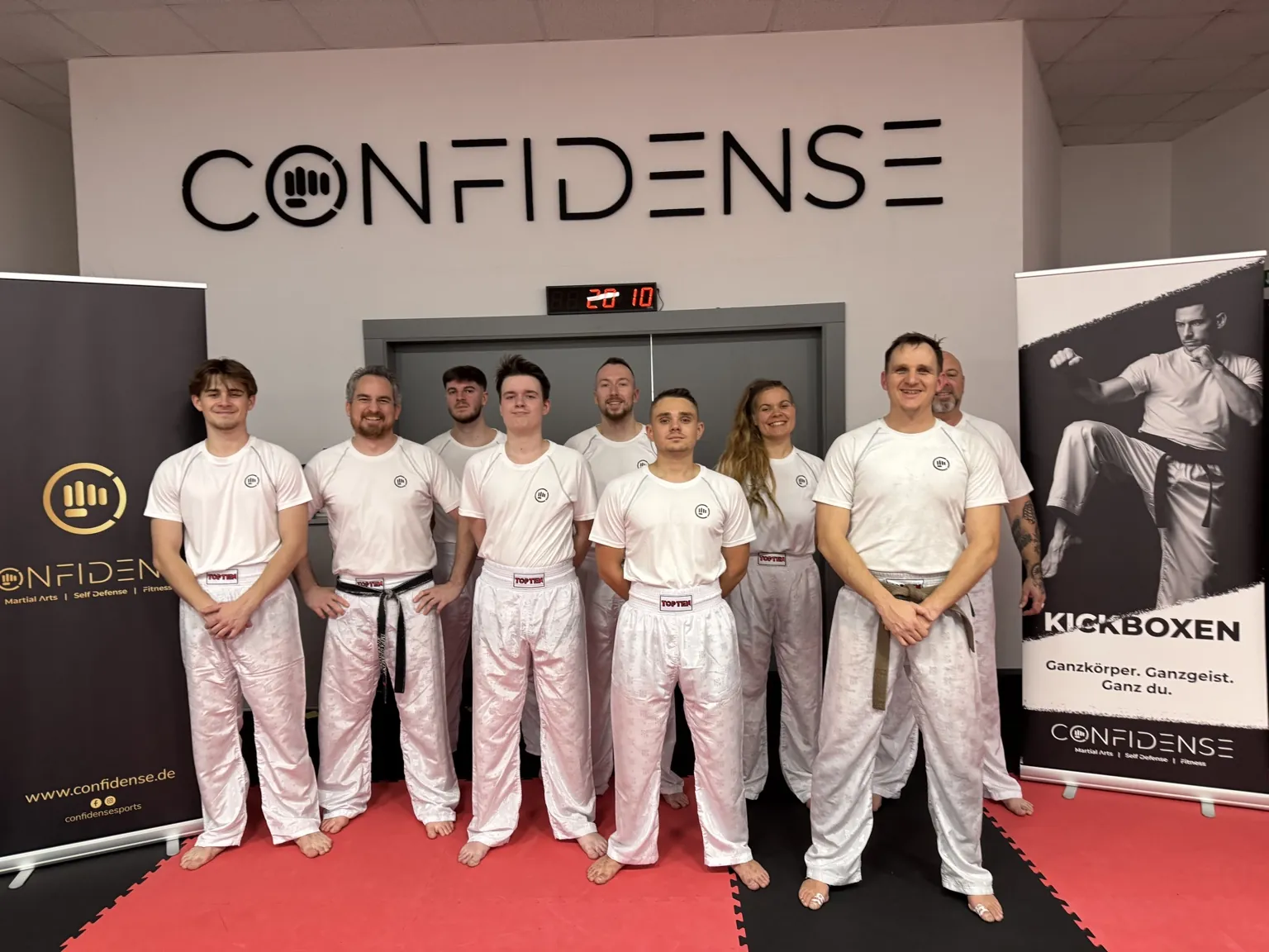 Große Gruppe Erwachsener in weißer Kickbox-Trainingskleidung steht gemeinsam im CONFIDENSE-Studio.