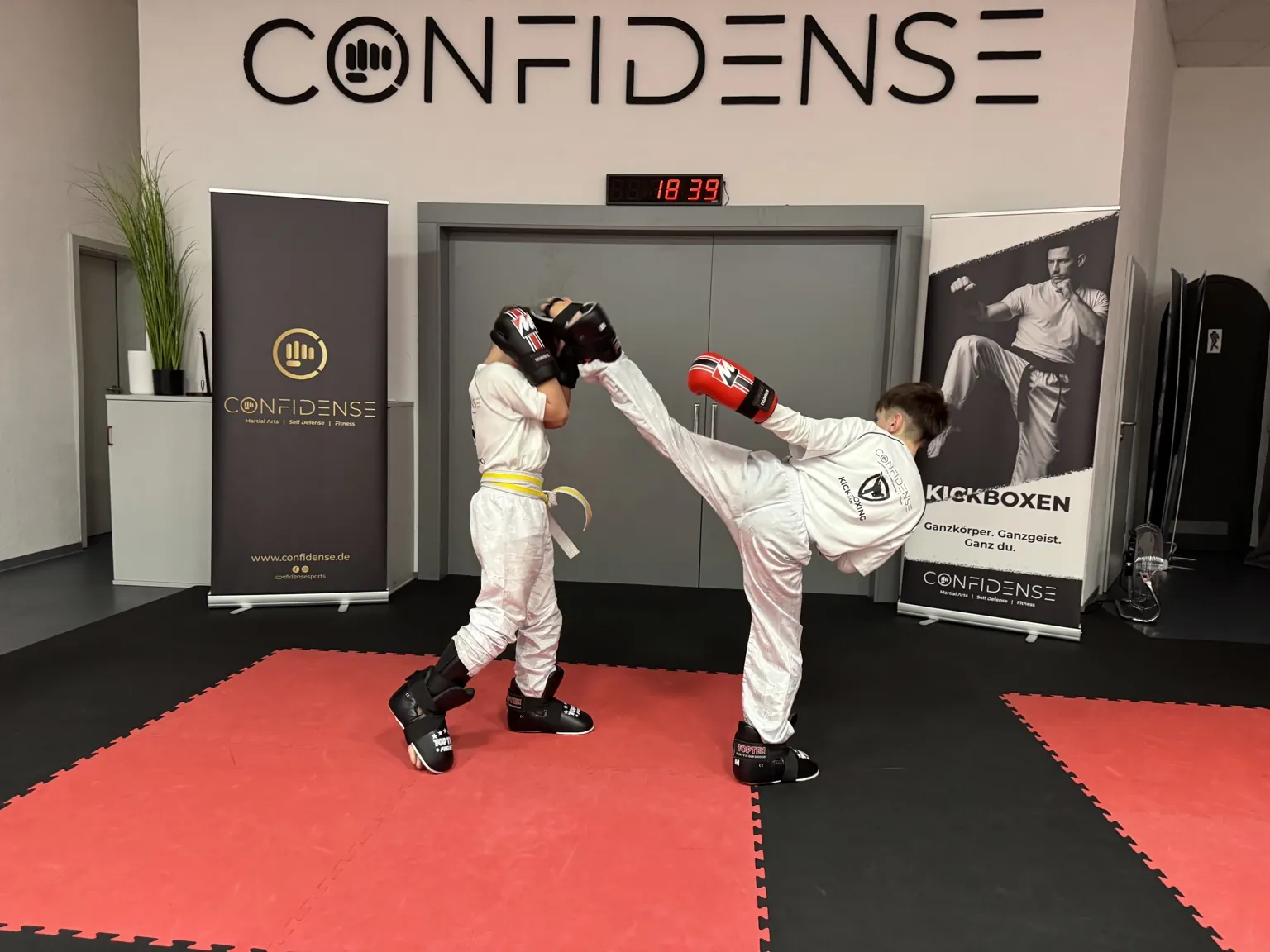 Zwei junge Kickboxer trainieren im CONFIDENSE-Studio einen hohen Tritt mit Schutzausrüstung.